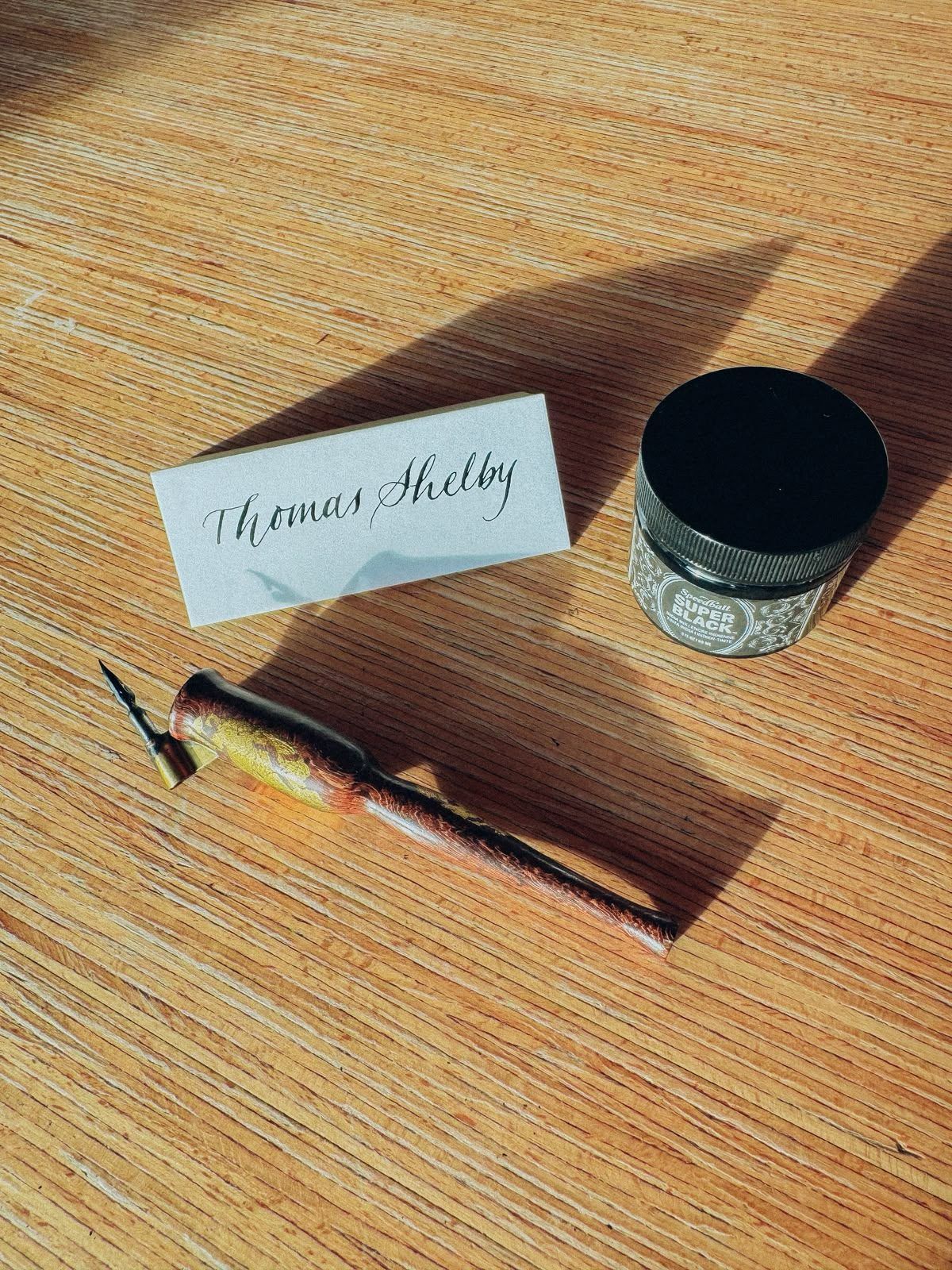 Marque-place calligraphié « Thomas Shelby », à côté d'une plume et d'un pot d'encre